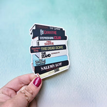 Load image into Gallery viewer, Hand holding a stack of books with various titles against a light blue background
