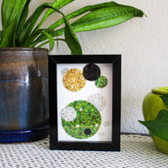 Decorative framed piece with colored stones on a table with plants in the background