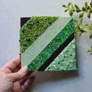 Hand holding a square piece of green textured material with a black stripe on a light gray background.