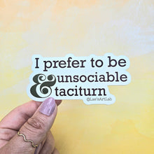 Load image into Gallery viewer, Hand holding a sticker with text 'I prefer to be unsociable &amp; taciturn' against a watercolor background.
