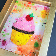 A decorative bead art piece resembling a strawberry chocolate cupcake with colorful seed beads and crystals on a gold-colored shadow frame.