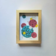 A framed geometric bead art piece featuring various colored beads arranged in a pattern, set in a gold shadow frame.