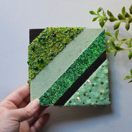 A square-shaped modern wall art piece with green stripes and decorated with glass seed and artisan beads, and Swarovski crystals, held by a hand against a neutral background.